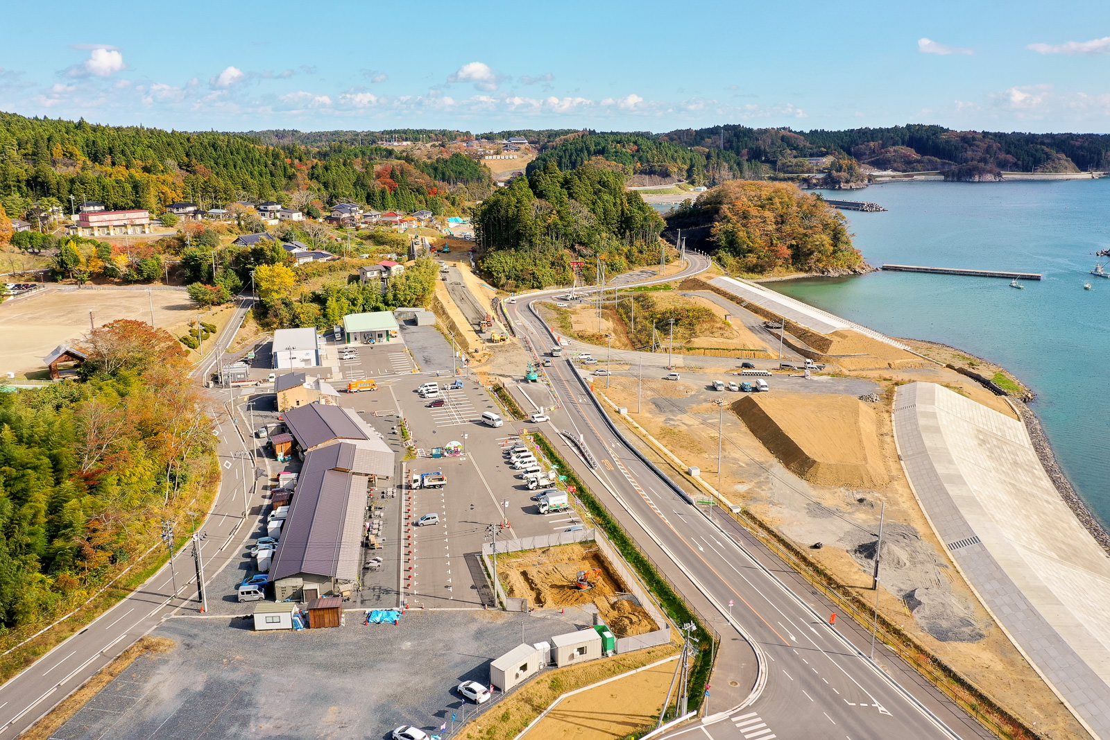 複数の建物や車両、工事機材が並ぶ広い駐車場を中心に、道路や木々、丘に囲まれた沿岸地域を空から見下ろした風景で、右側には護岸整備された海岸線と水面が広がる空撮の写真