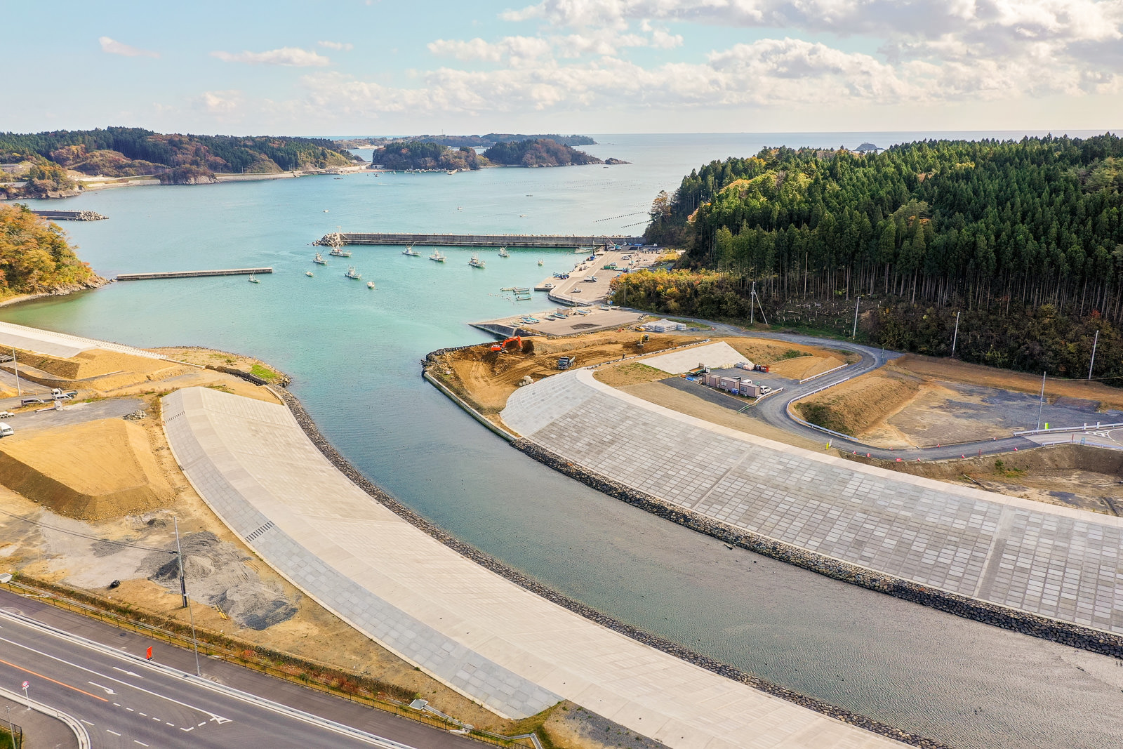 川が海へと流れ込む海岸地域を空から見下ろした風景で、川の両岸にはコンクリートの護岸が整備され、川沿いには道路が走り、海には小型の船がいくつか停泊し、片側には森林が広がり、背景には丘が見え、空には雲が浮かぶ空撮の写真