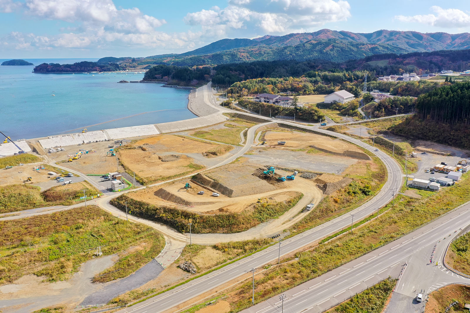海に面した地域で建設作業が進められている様子を空から捉えた風景で、複数の建設車両や機材が点在し、周囲には道路が整備され、片側には海が広がり、背景には緑に覆われた丘や山々が見える沿岸工事現場の写真
