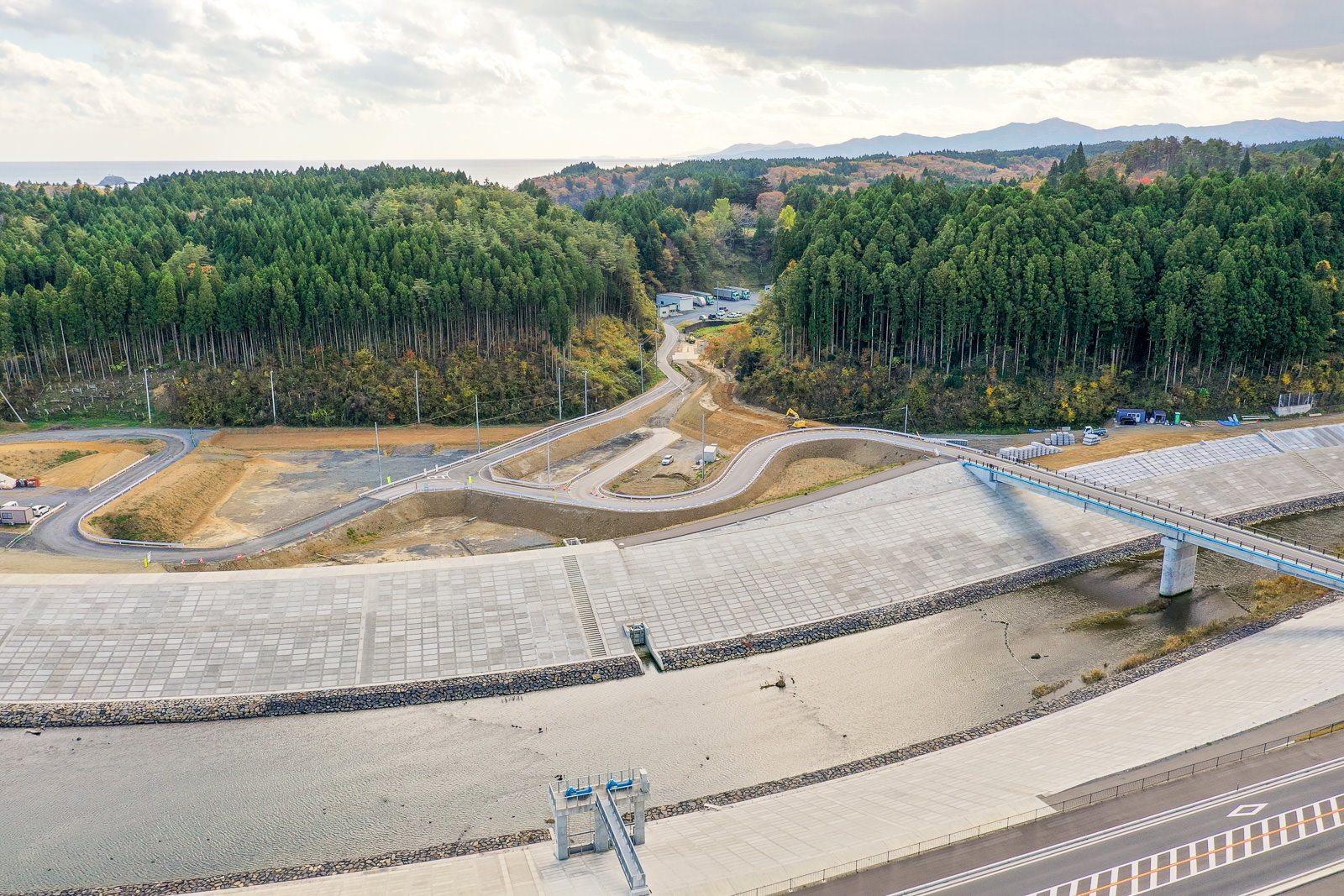 手前に水が流れる大きなコンクリート構造物があり、ダムまたは治水施設と思われる風景で、奥には曲がりくねった道路が両側の密集した森林の中を通り抜け、さらに遠くには丘が広がり、空には雲が浮かぶ空撮の写真