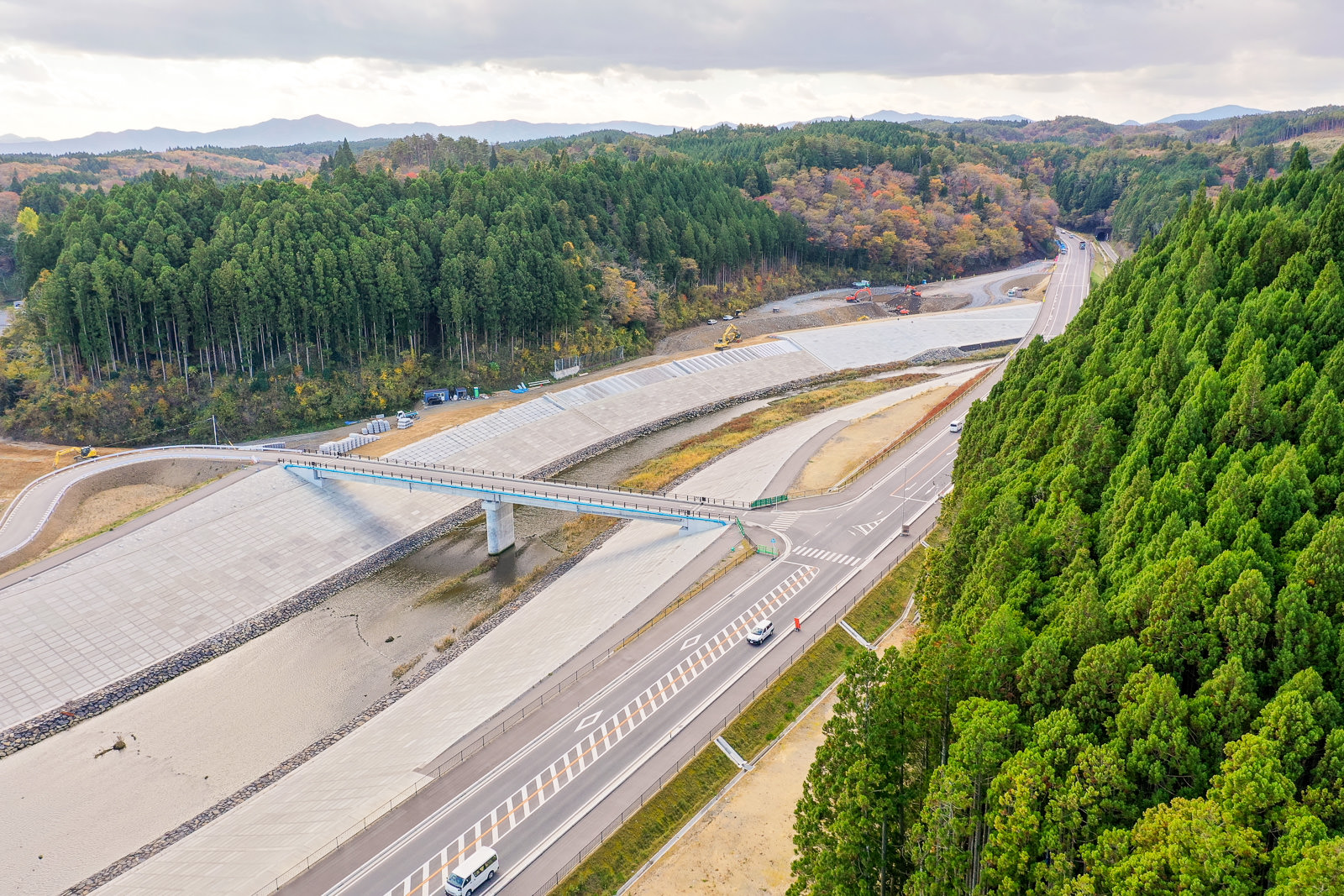 川または運河に架かる橋と、その両側に広がる密集した森林を空から見下ろした風景で、川沿いには車が走る道路があり、背景には丘や山々が広がり、空には雲が浮かぶ空撮の写真