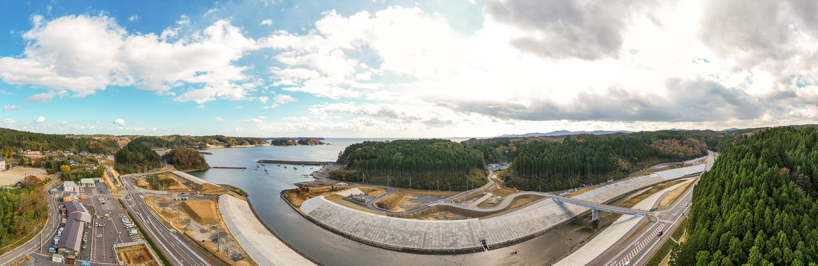 川が海へと流れ込む海岸地域を広く見渡した風景で、川には複数の橋が架かり、周囲には道路や建物、密集した森林が広がり、空には雲と青空が見える空撮の写真