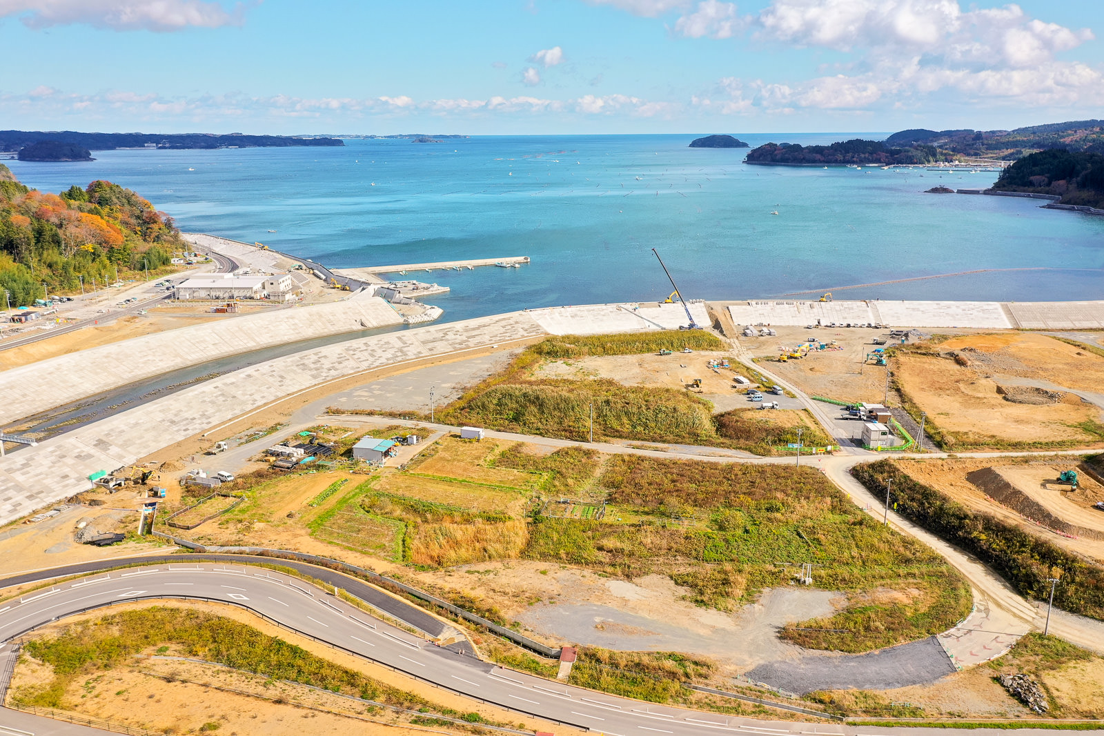 背景に広がる大きな水域と、遠くに点在する小さな島々を望みながら、手前には道路や芝生、建設機材が配置された開発中の沿岸地域が広がり、下部には水辺へと続く湾曲した道路が目立つ晴天の海辺の写真
