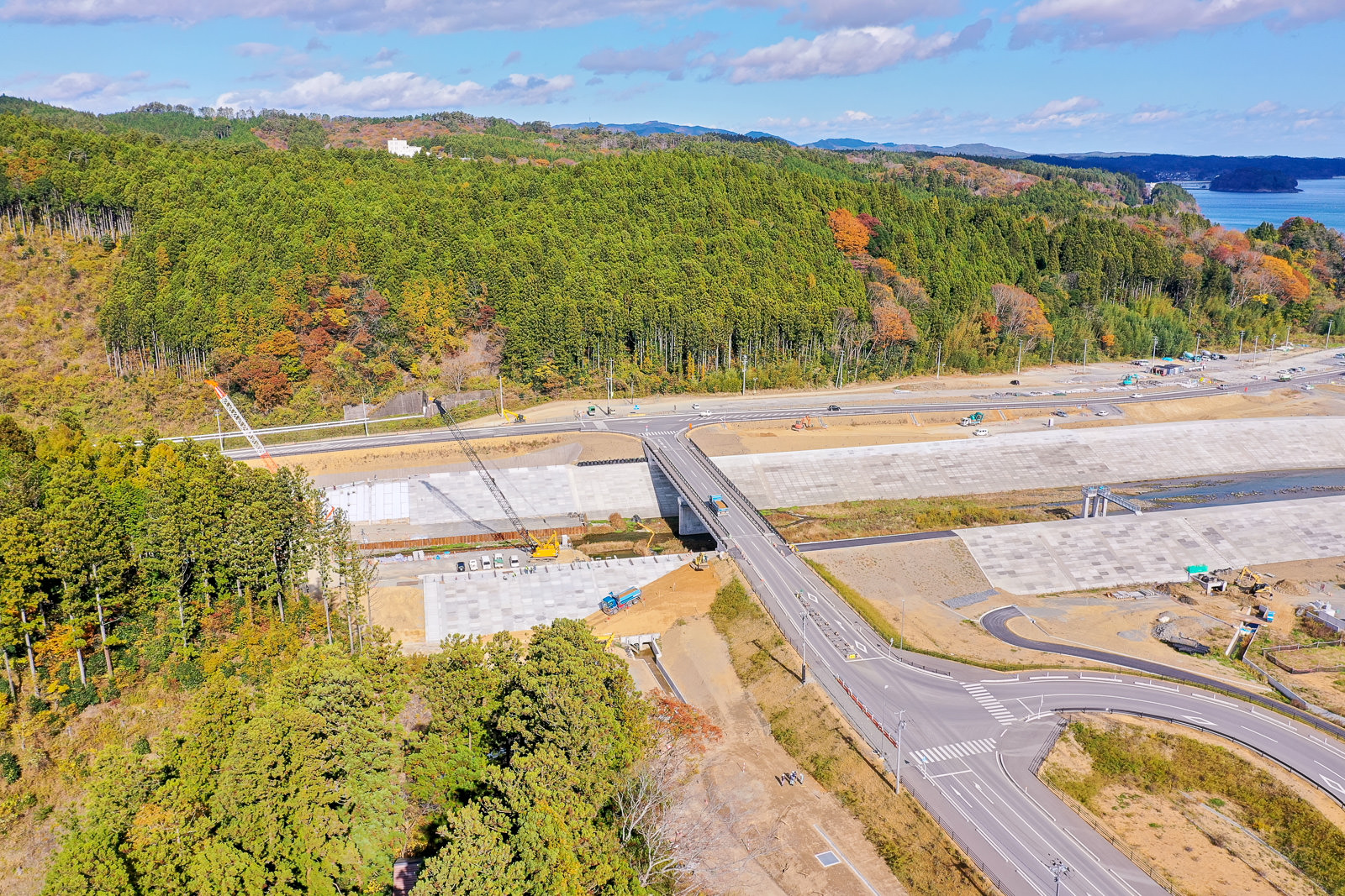 道路が通る工事現場を空から捉えた風景で、道路の上には橋が架かり、周囲は密集した森林に囲まれ、空には青空と雲が広がる自然の中の建設現場の写真