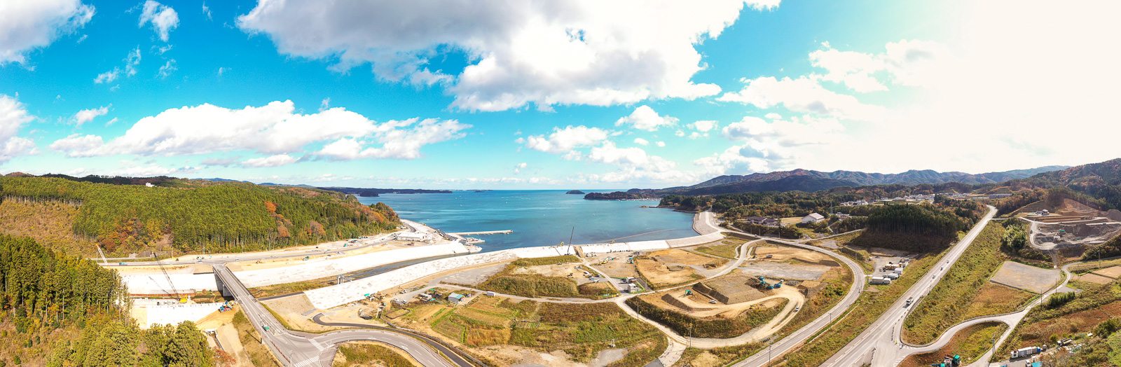 中央に水域が広がり、左側には丘と森林が広がる自然の風景が見られ、右側には道路や建設現場、建物が点在する開発が進む地域が広がり、空には青空と雲が混在する沿岸地域の写真