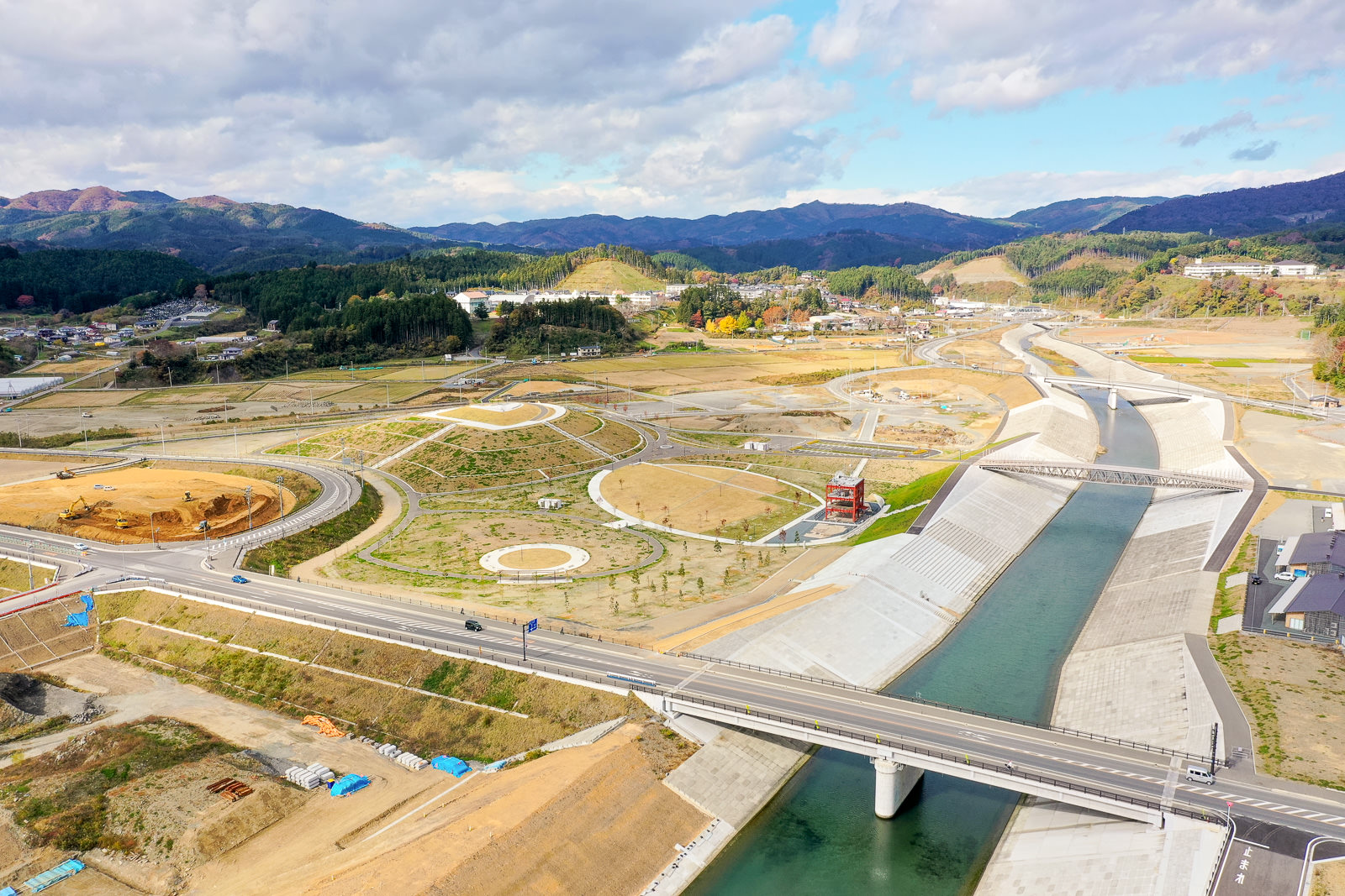コンクリートで整備された川の両岸に橋が架かり、その周囲には円形に植栽された緑地や道路が広がり、遠くには丘や山々が見える空撮風景の写真