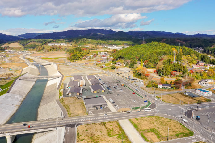 コンクリートで護岸された川または運河に橋が架かり、その周囲に建物や駐車場が広がり、手前には交差する道路と車が見え、背景には木々に覆われた丘と点在する建物が広がる都市近郊の風景の写真
