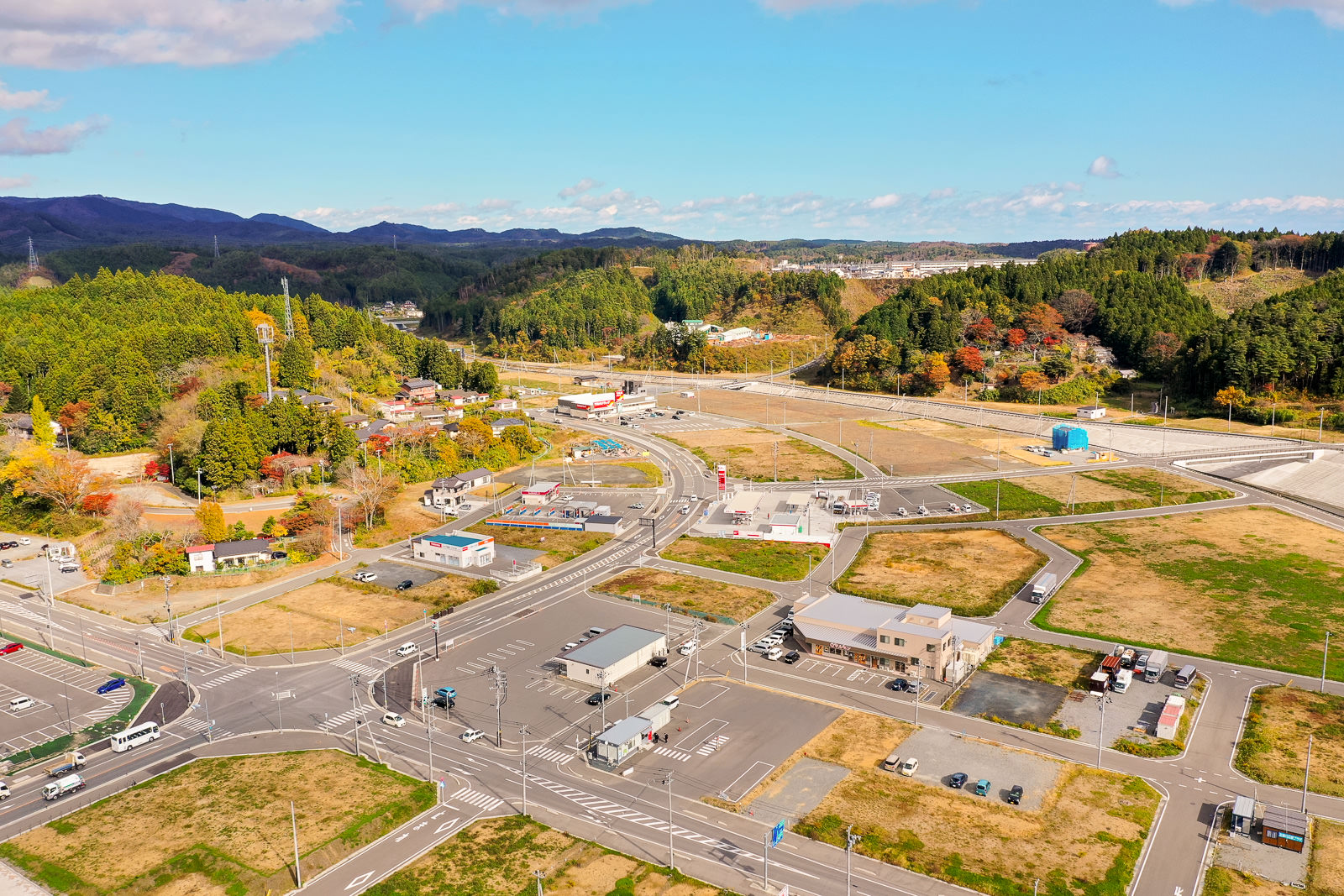 道路網と建物、広い畑が広がる農村地帯を空から見下ろした風景で、交差点や駐車場が点在し、周囲には緑の丘や森林が広がり、所々に住宅や構造物が見える空撮の写真