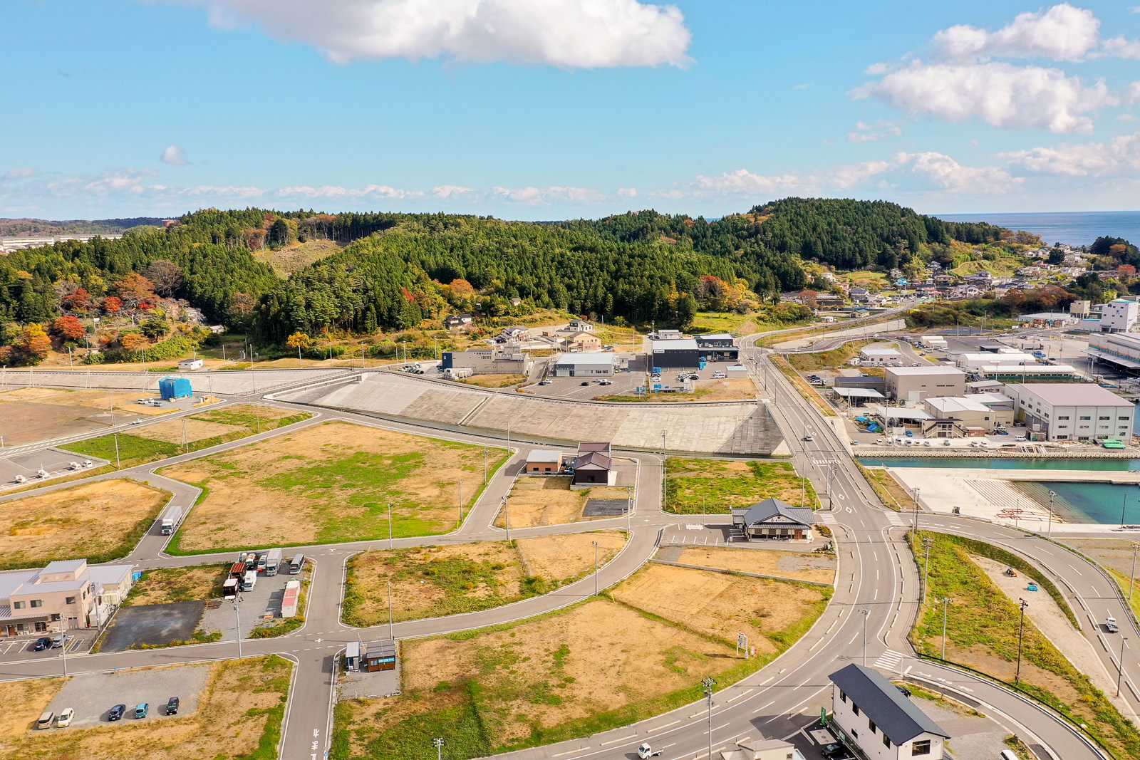 住宅と工場が混在する地域を空から見下ろした風景で、交差する道路沿いに家屋や車両が点在し、緑の畑や茶色い土地が広がり、背景には森林に覆われた丘とその先に海が見える空撮の写真
