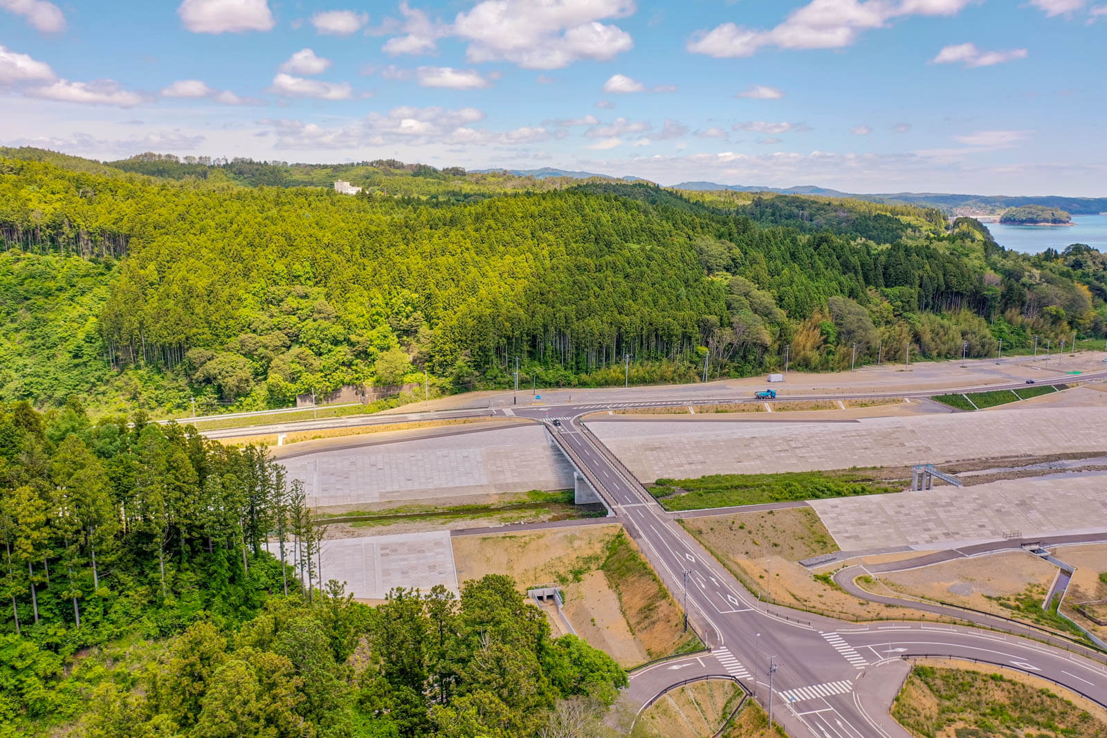 青空の下、濃淡の緑が広がる森林と、その手前に整備された広い護岸や交差点、道路が走る風景を上空から撮影した写真