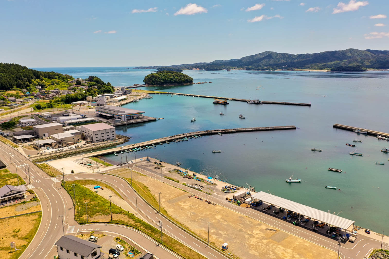 手前には道路網と広場が整備され、奥には複数の漁業関連施設や倉庫が並んでおり、港は防波堤で囲まれ、内側には多くの小型漁船が停泊している写真
