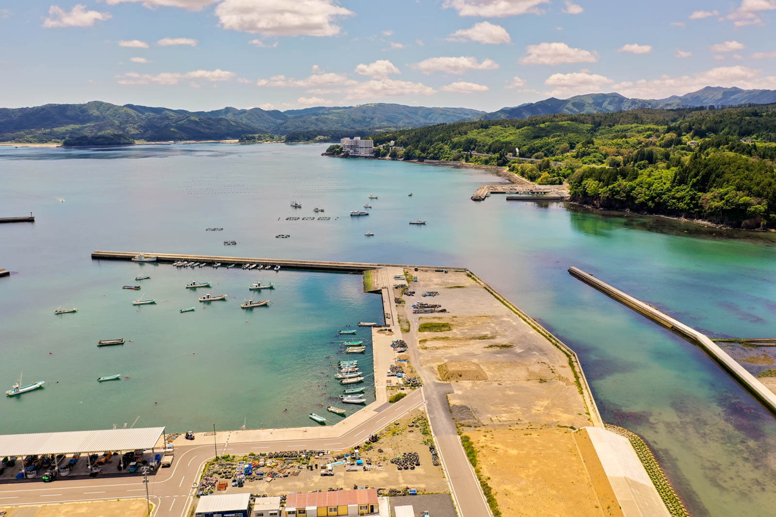 手前には漁船が並ぶ小さな港と、防波堤で囲まれたエリアがあり岸壁の背後には広い空き地や資材置き場があり、港湾設備として利用されている様子を写した写真