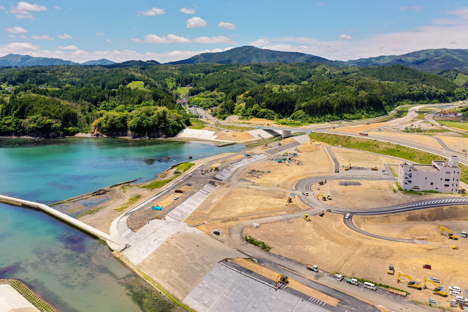 手前には新しく整備された堤防や護岸が見え、海と陸を分ける防潮設備がしっかり構築されており、右側には工事中の道路や重機が点在し、復興や再開発の過程である写真