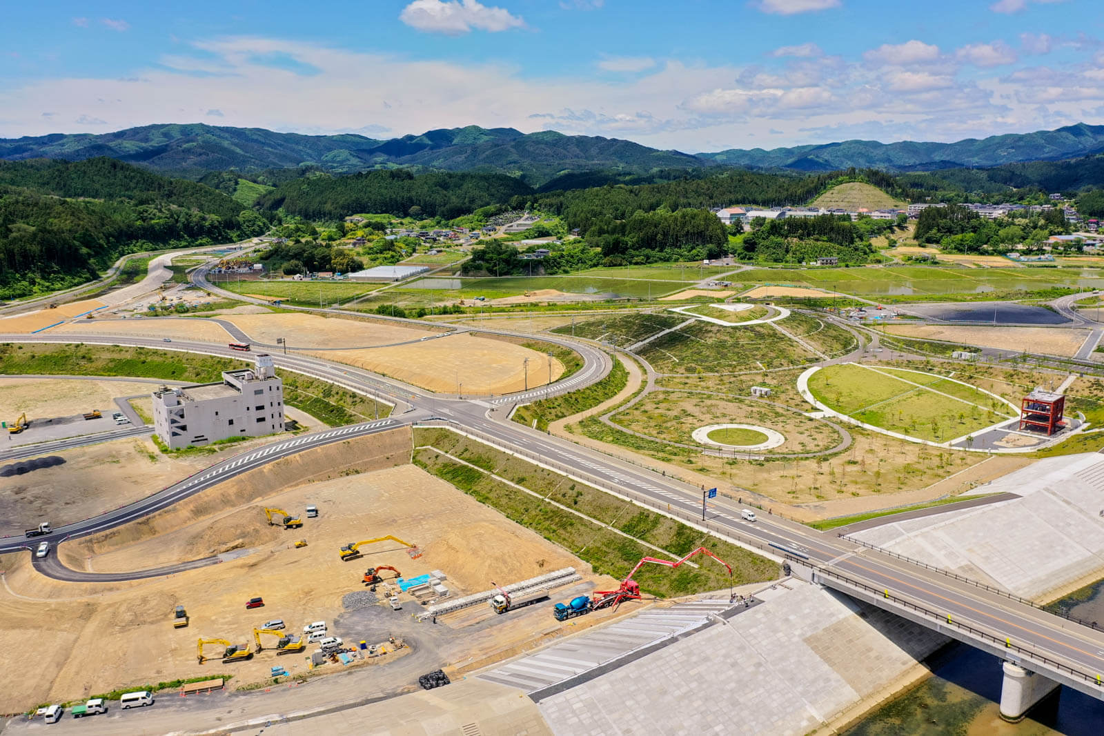 右側には円形や曲線を多用した整備済みの緑地空間があり、左側には白い建物があり、その周囲や手前の区画では工事車両が稼働しており、道路や護岸の造成工事が進行している様子を写した写真