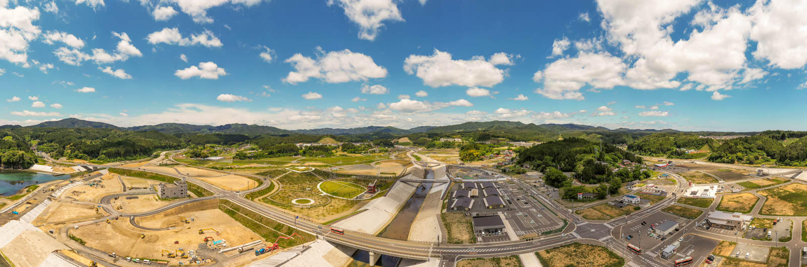中央には大きな川とそれをまたぐ橋、新しい道路や造成地、そして右側には整然と並んだ建物群（商業施設や市場のような建物）が見え、奥には山々が連なり、全体的に復興と再開発が進んでいる様子のパノラマ写真