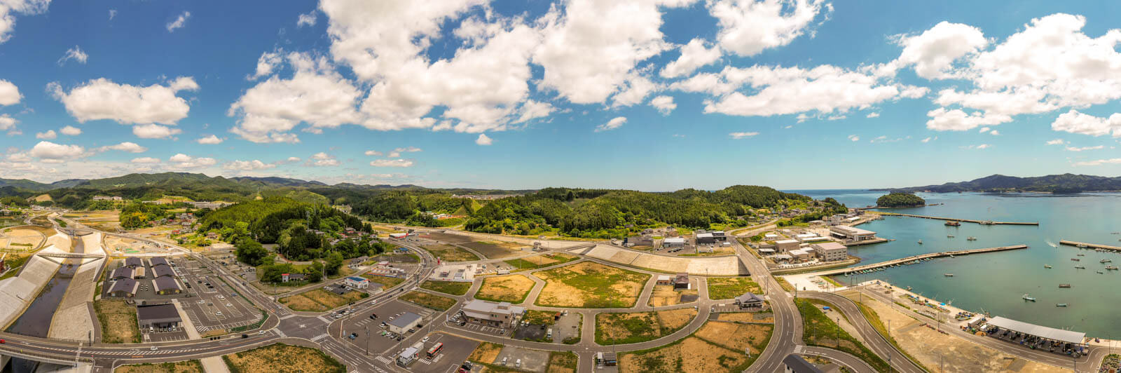 左側には堤防と川、整備された駐車場や施設群が見え、中央から右にかけては緑豊かな丘と、その向こうに広がる海、港湾施設、そして停泊する多くの船が映っているパノラマ写真