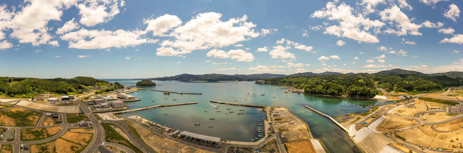 穏やかな入り江には複数の防波堤が伸び、漁船や小型船が停泊している港湾とその周辺地域を広角で捉えたパノラマ写真