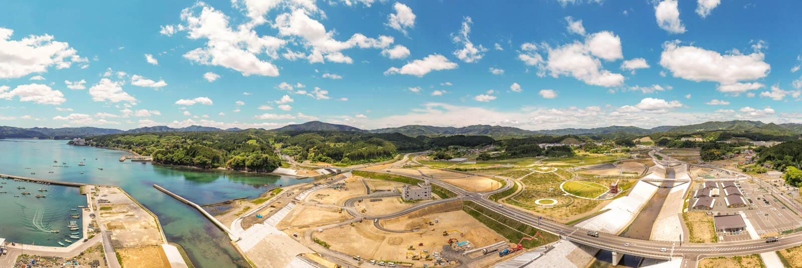 左側には穏やかな湾と漁港、防波堤、そして停泊する漁船が見え、その隣には港湾施設があるパノラマ写真