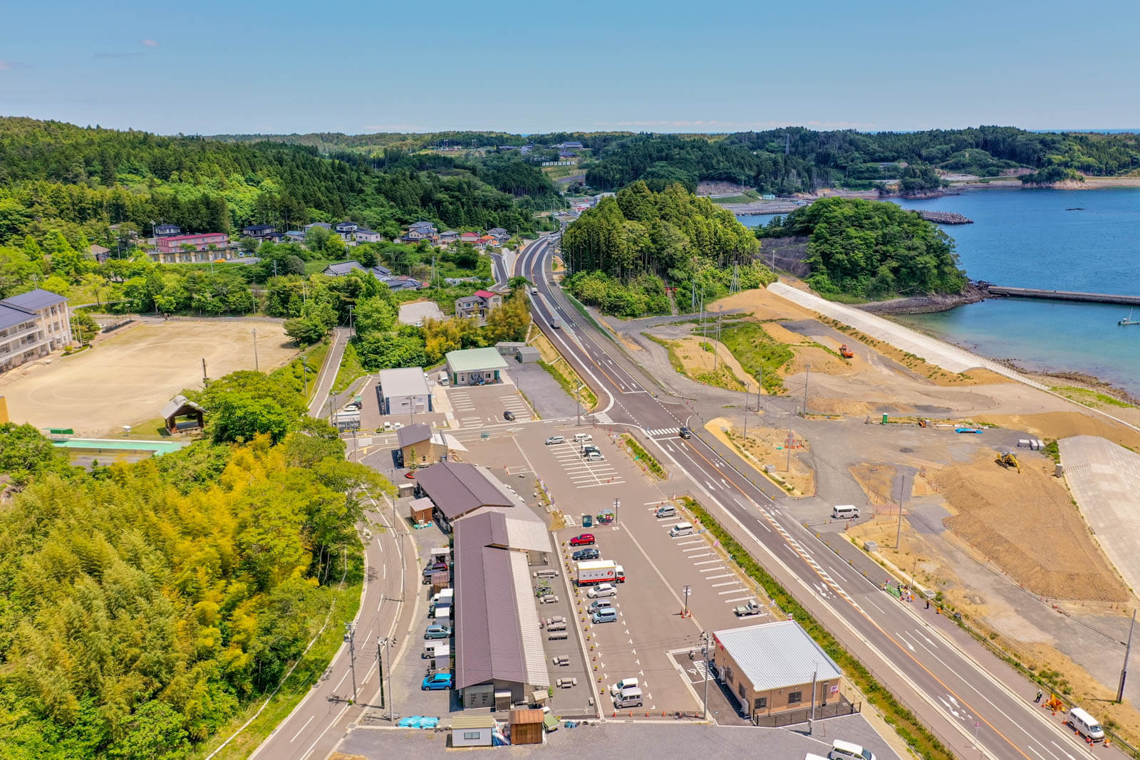 手前には駐車場と複数の建物が並んでおり、左側には学校のグラウンドが見え、その奥には住宅が点在している写真