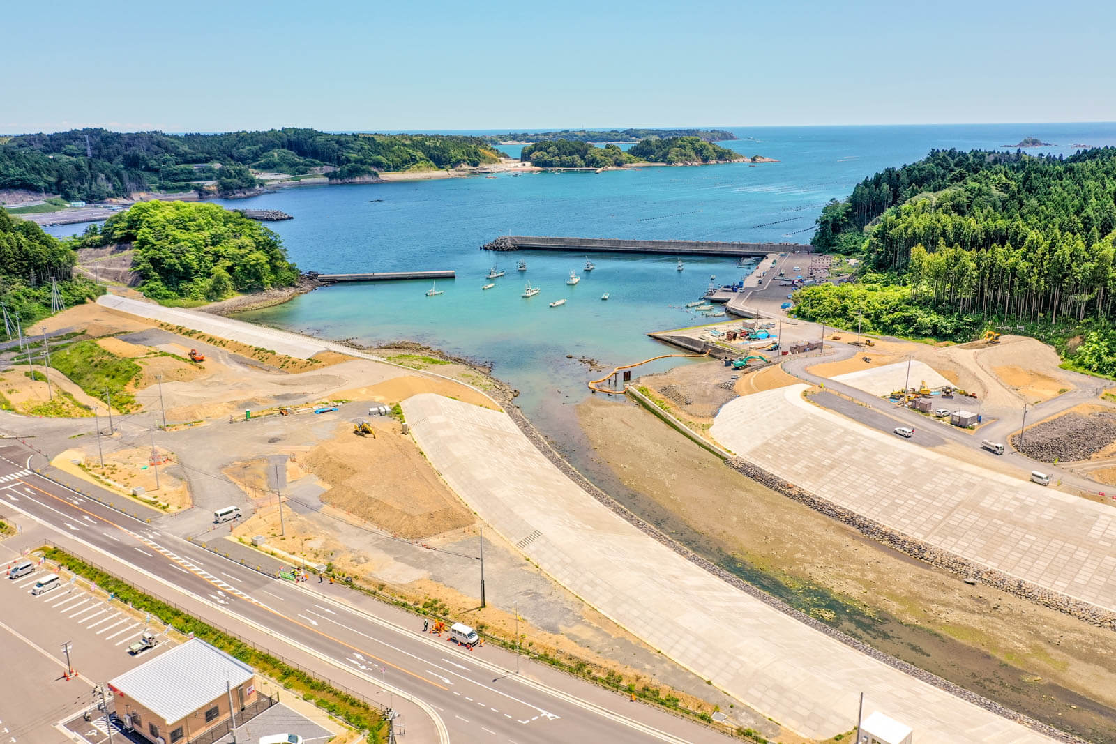海面には小型漁船が浮かび、背後には森林に覆われた岬や島が見え小さな湾と港を上空から撮影したもので、防波堤に囲まれた穏やかな海域が広がっている写真