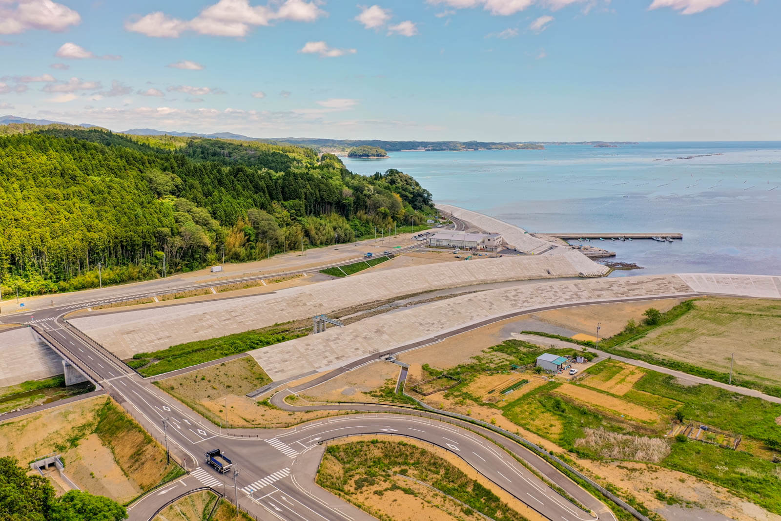 青い海と港を背景に、防潮堤と護岸工事が整備された海岸沿いの道路や交差点、周辺の緑地や農地が広がる風景を空撮した写真