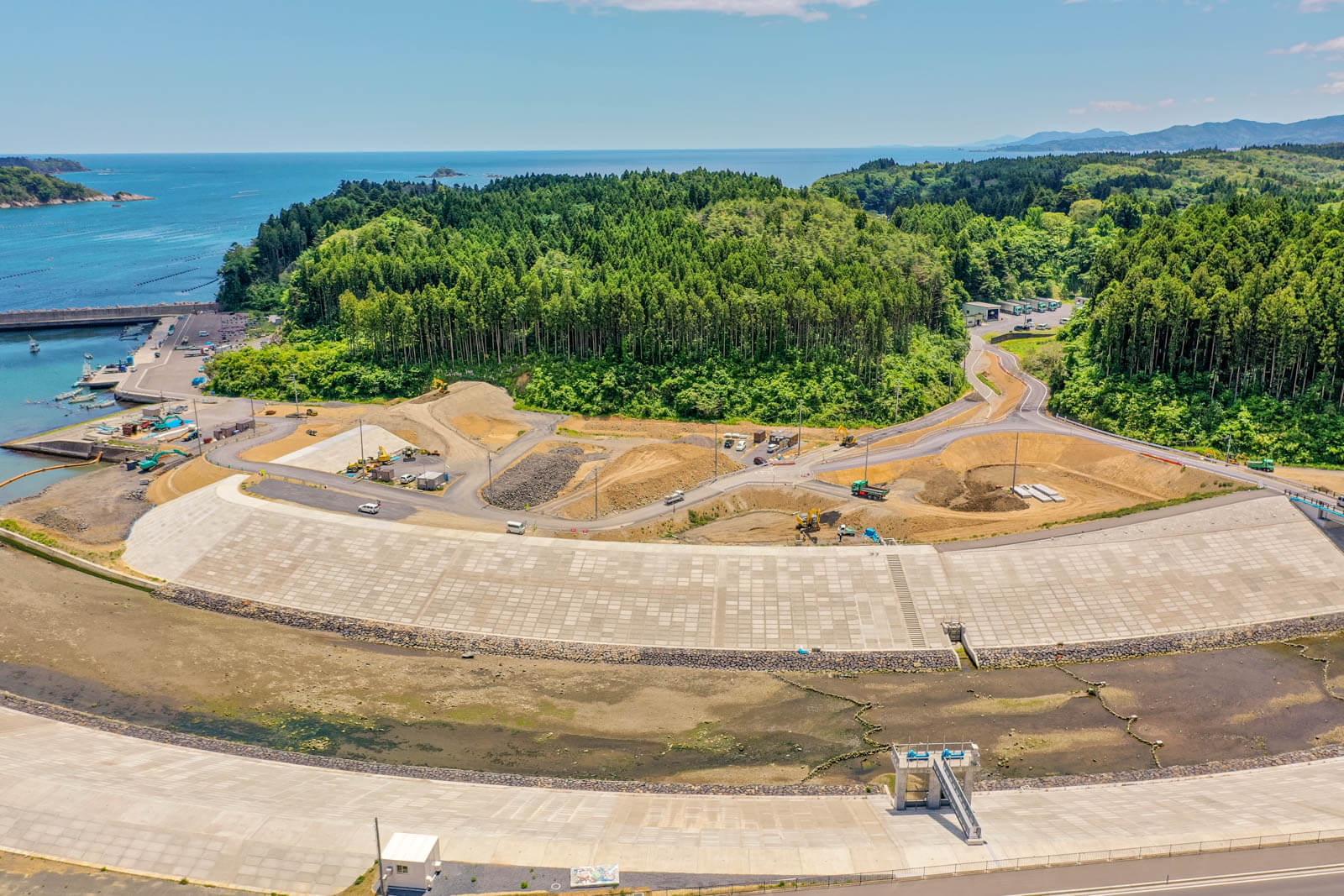 手前には高さのあるコンクリート製の防潮壁と、その中央に設置された水門施設が見え奥には森林に覆われた小高い丘があり、その手前の造成地では複数の建設機械やトラックが作業をしている様子を写した写真