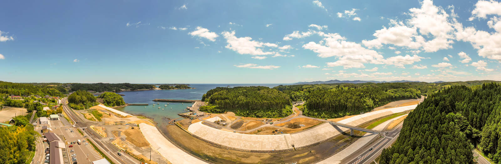 中央には防潮堤と護岸がしっかり整備され、川の流れを海へ導いて右側は山が迫り、濃い緑の森林が広がる一方、左側には港と防波堤があり、小型船舶が係留されている様子を写した写真