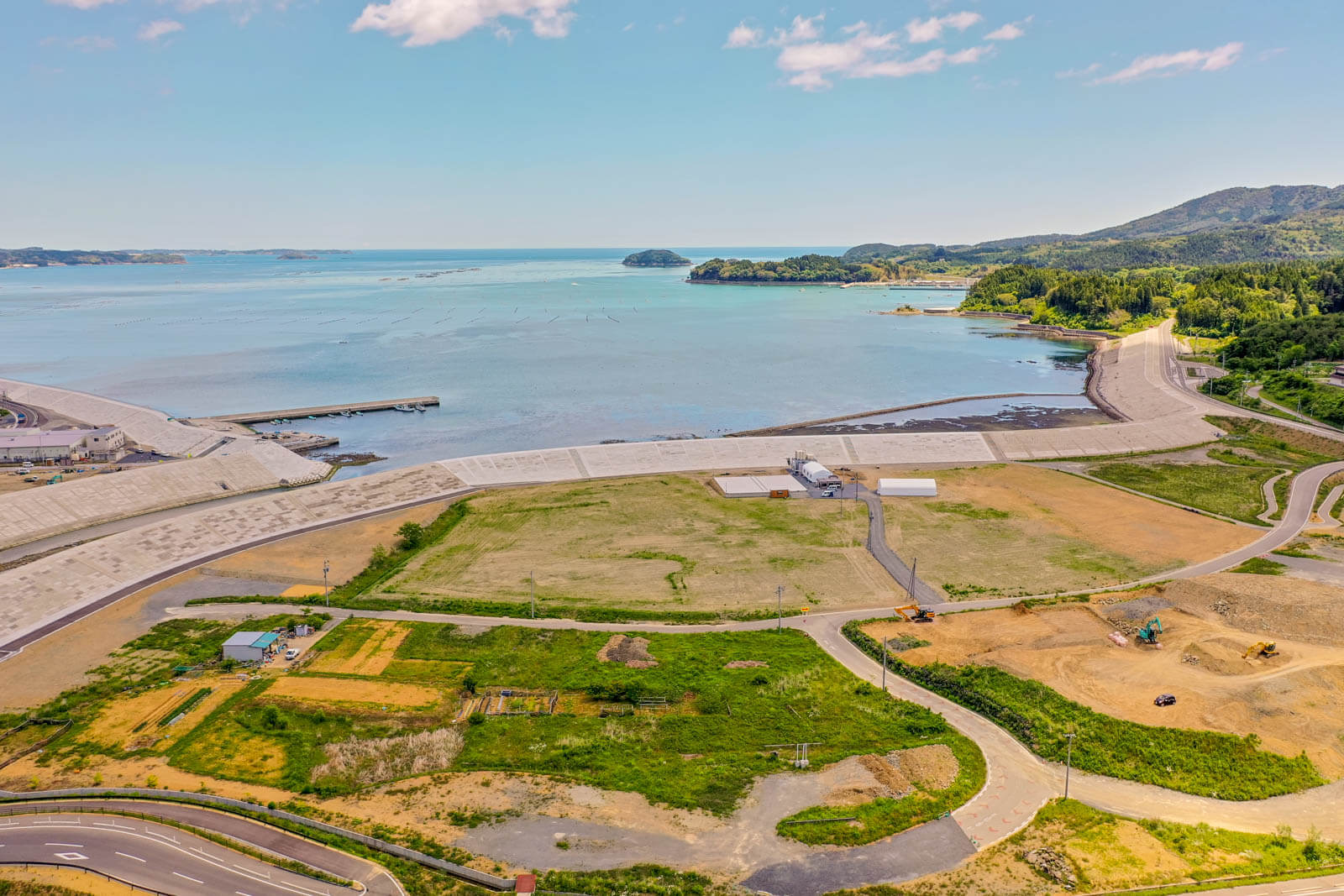 青い海と防潮堤、港湾施設を背景に、広い造成地や緑地、重機が稼働する工事現場が広がる海沿いの風景を空撮した写真