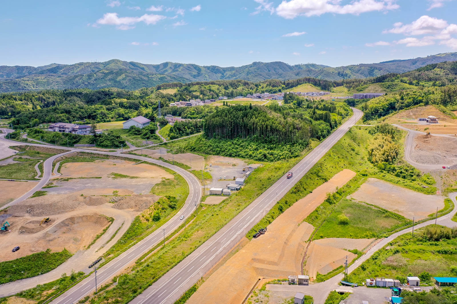 山々を背景に、新しい道路や造成地、工事中のエリア、点在する建物や住宅が広がる復興途中の町並みを空撮した写真です