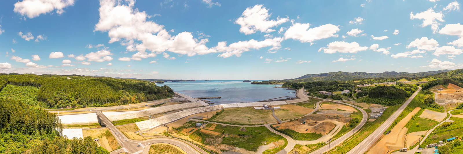 青空の下、緑豊かな山々と防潮堤で囲まれた湾岸、港や護岸工事の進む沿岸部と周辺の道路や造成地が一望できるパノラマ空撮写真