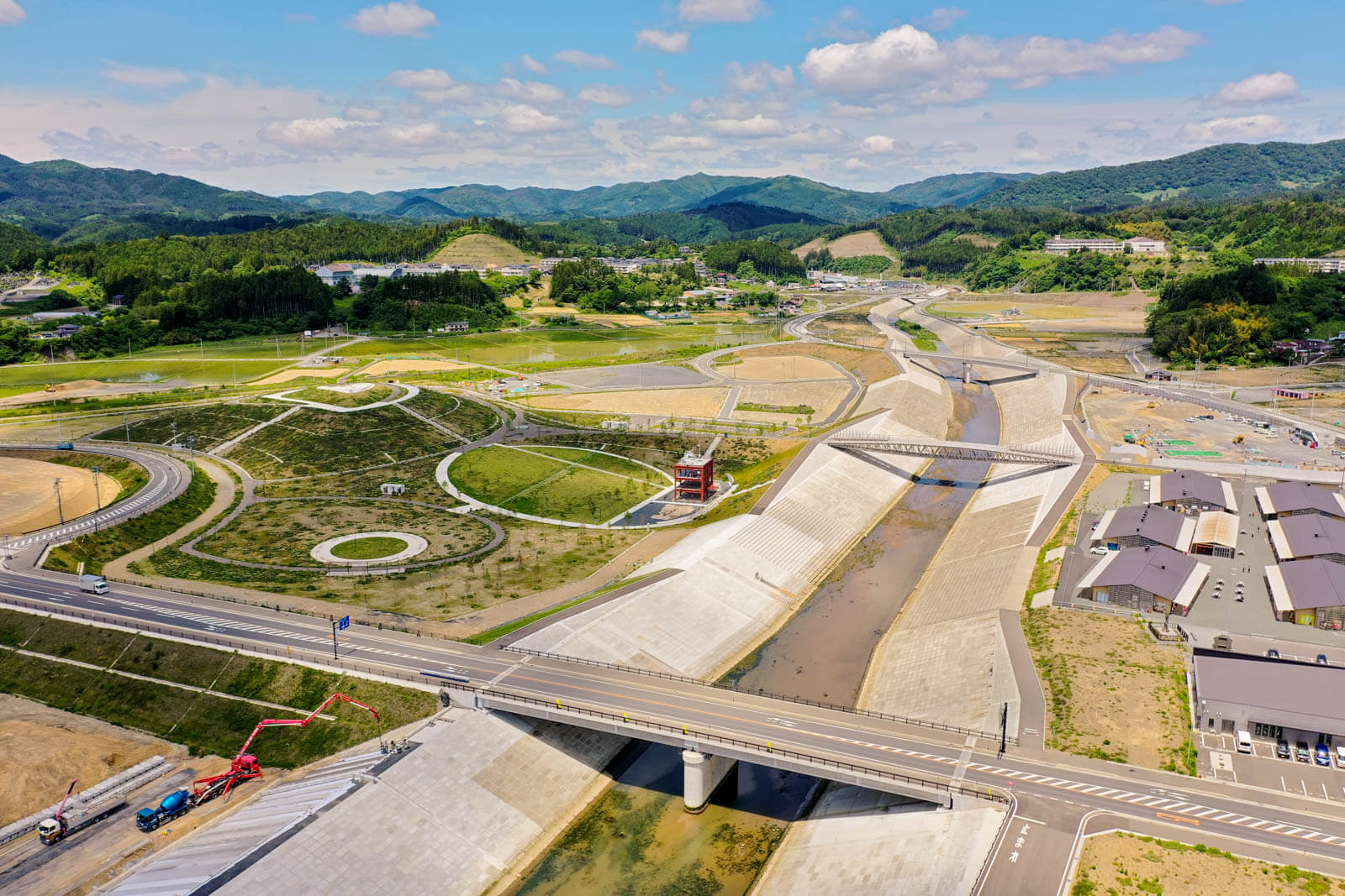 中央には広い川と堤防、そして複数の橋が見え、その横には新しい道路や円形の公園のような緑地スペースがあり、右下には整然とした商業施設の建物群が並び、左下では大型クレーン車やコンクリートミキサー車が作業中の写真