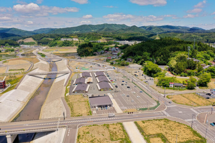 青空の下、山々を背景に整備された河川護岸と橋、広い駐車場を備えた複数の平屋建物が並ぶ施設と周辺の道路が見渡せる空撮写真