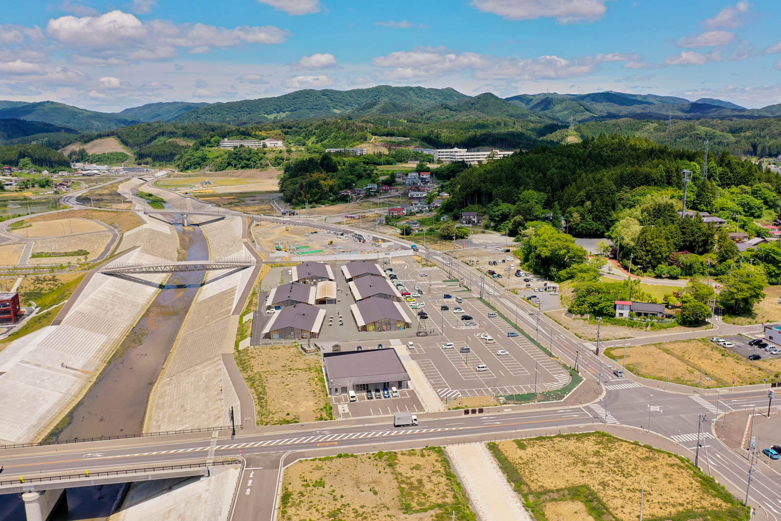 中央には川と両岸の高い堤防、その間に架かる歩道橋が見え、川の右側には整然と並ぶ複数の建物群と大きな駐車場がある写真