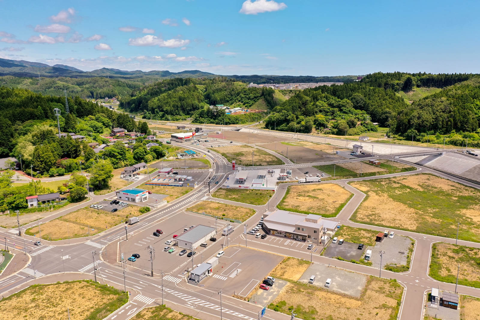 道路網と整備された区画がはっきり見え、手前には駐車場付きの建物やその背後には緑豊かな丘と住宅地が広がっている写真