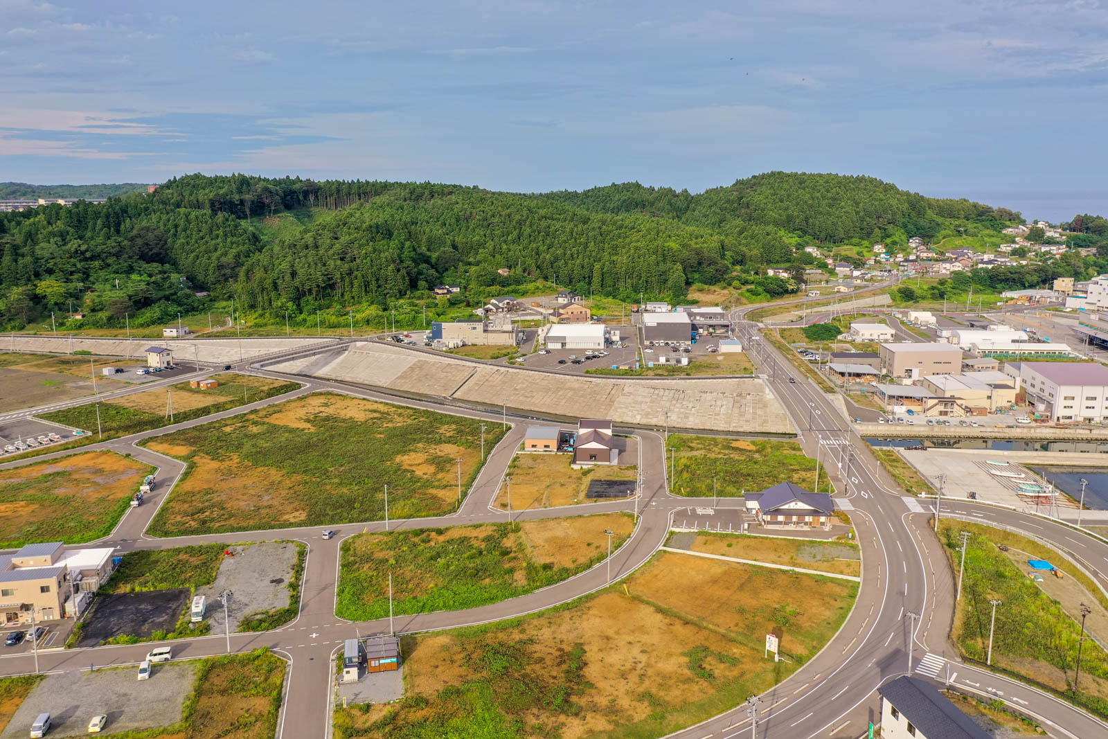 複数の道路と建物、緑地が広がる郊外の地域を空から見下ろした風景で、建物のある区画と空き地が混在し、背景には緑豊かな丘や木々が広がる晴れた日の写真