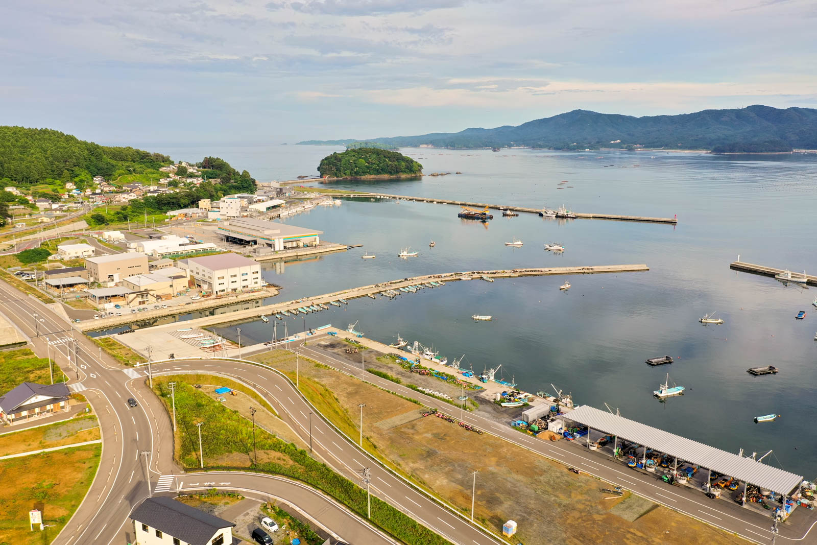 港には複数の船が停泊しており、岸沿いには建物が並び、周囲には道路と緑地が広がり、背景には丘が連なり、遠くには島が浮かぶ穏やかな海辺の町の写真