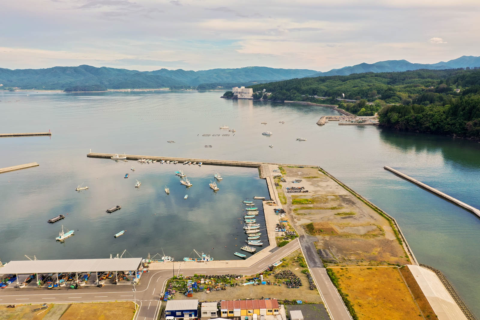 港には複数の船が停泊しており、周囲には建物や道路、緑地が広がり、背景には山々が連なる沿岸地域を空から見た写真