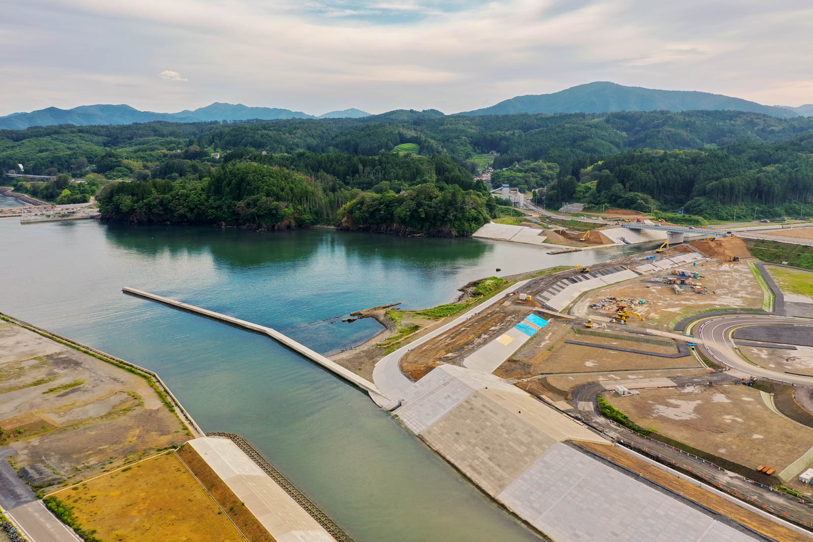 海に面した地域を空から見下ろした風景で、海岸線にはコンクリートの防潮堤が設けられ、周囲には木々に覆われた丘や一部開発中の土地が広がる沿岸地域の写真