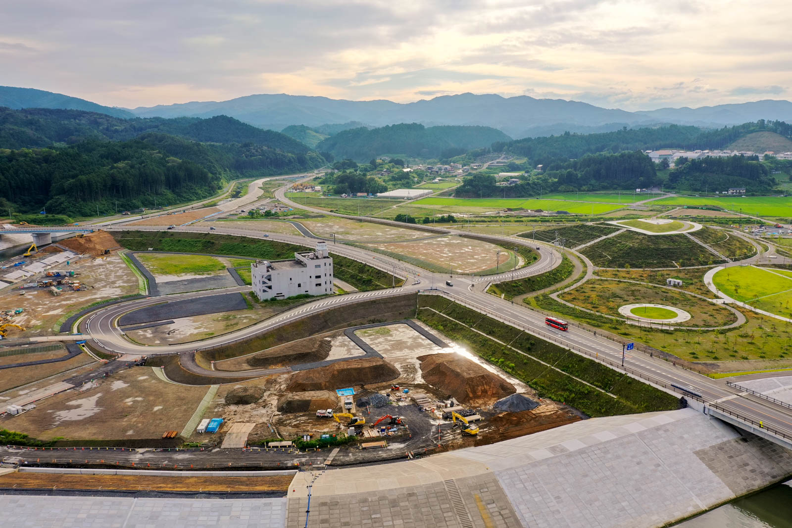複雑に交差する道路や高速道路が広がり、手前には建設機材が並ぶ工事現場があり、中央には白い建物が道路に囲まれ、背景には緑の畑や木々、山々が広がる曇り空の下の風景の写真