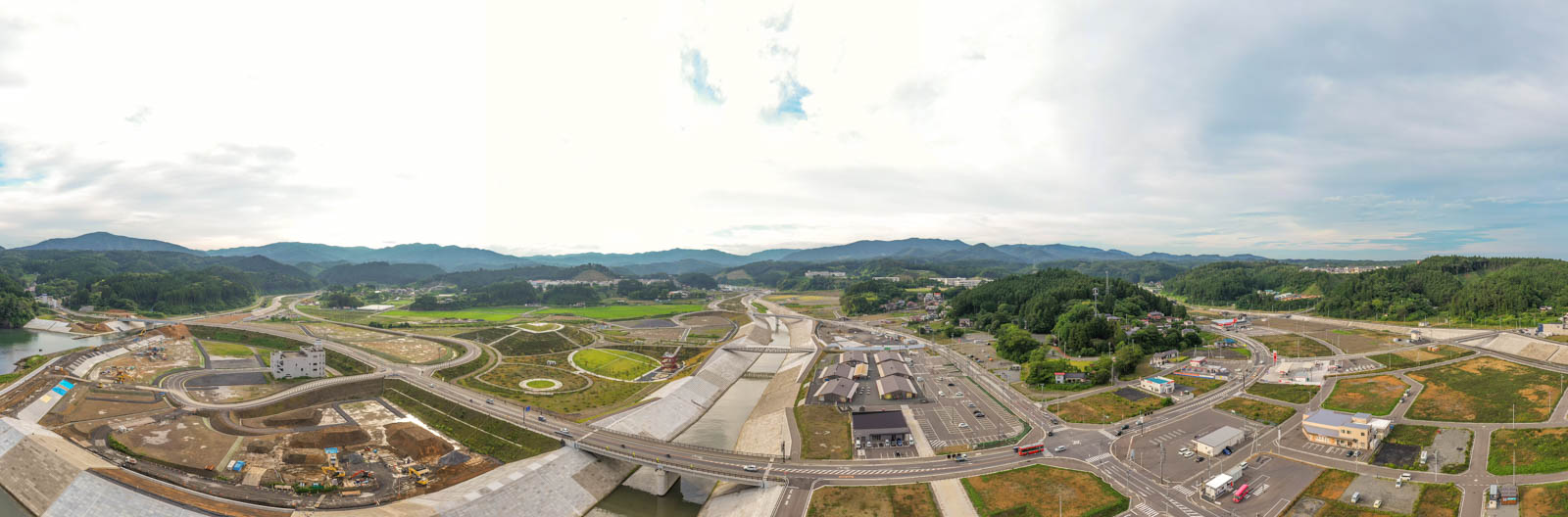 手前に道路や橋、建物などのインフラが広がり、奥には緑の畑や丘、山々が連なり、空には青空と雲が混ざる田舎の広域風景の写真