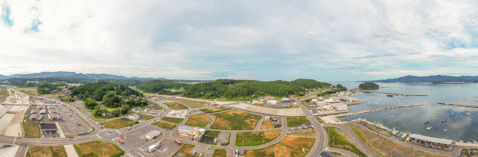 右側に港があり、船が停泊していて、周囲には建物や道路、緑地が広がり、背景には緑に覆われた丘が連なる、曇り空の下の沿岸の町を空から見た写真