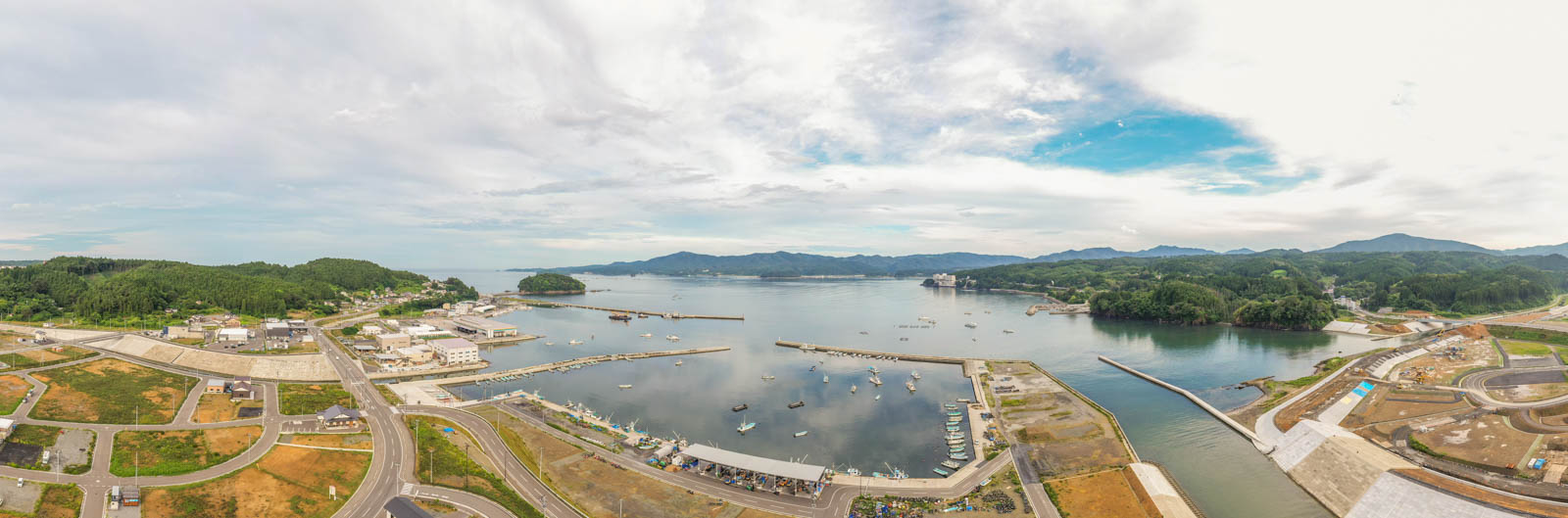 港には複数の船が停泊しており、その周囲に建物や道路などのインフラが広がり、背景には緑の丘が連なり、空には青空と雲が混ざる沿岸の町を空から見た写真