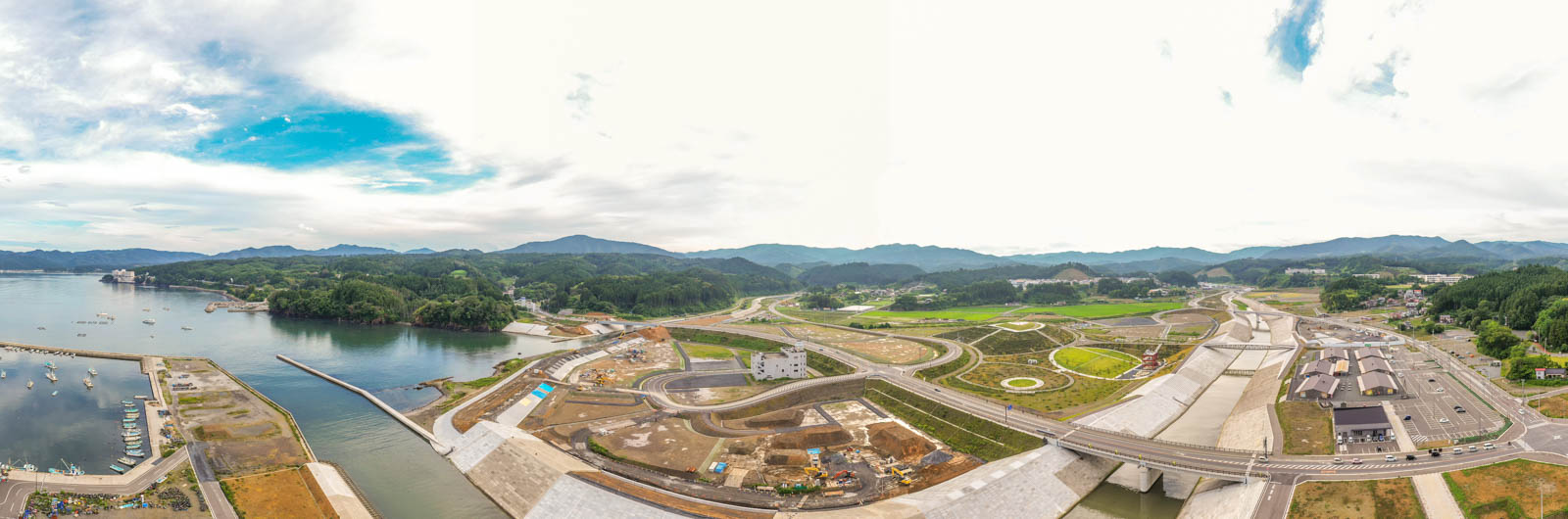 左側に海とマリーナが広がり、船が停泊していて、周囲には道路や橋、工事現場などのインフラが点在し、背景には緑の丘と山々が連なる曇り空の下の沿岸地域の写真