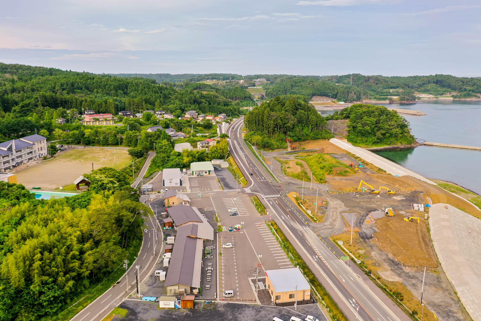 道路や建物、駐車場、工事現場が点在し、緑の森林や住宅地、海岸線を含む沿岸地域を空から見下ろした、晴れた昼間の風景の写真