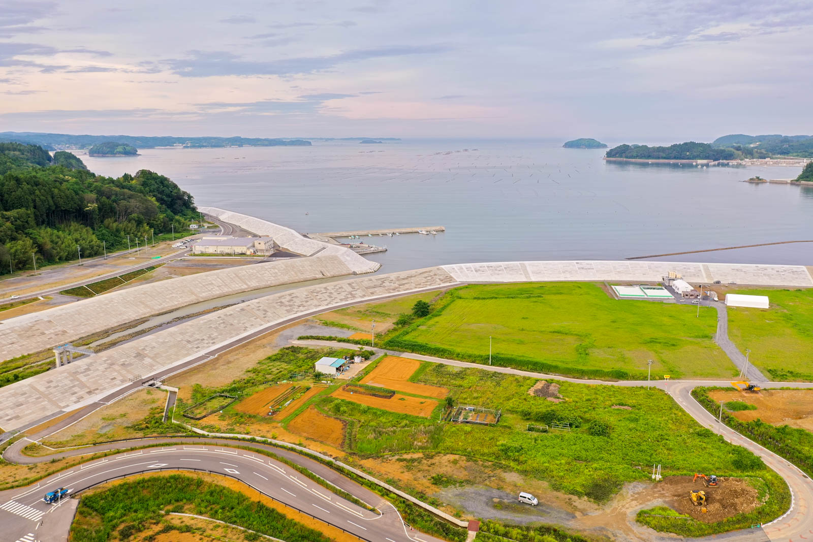 海沿いに設けられた防波堤と、その内側に広がる緑の畑や道路が見える空からの視点による沿岸地域の写真