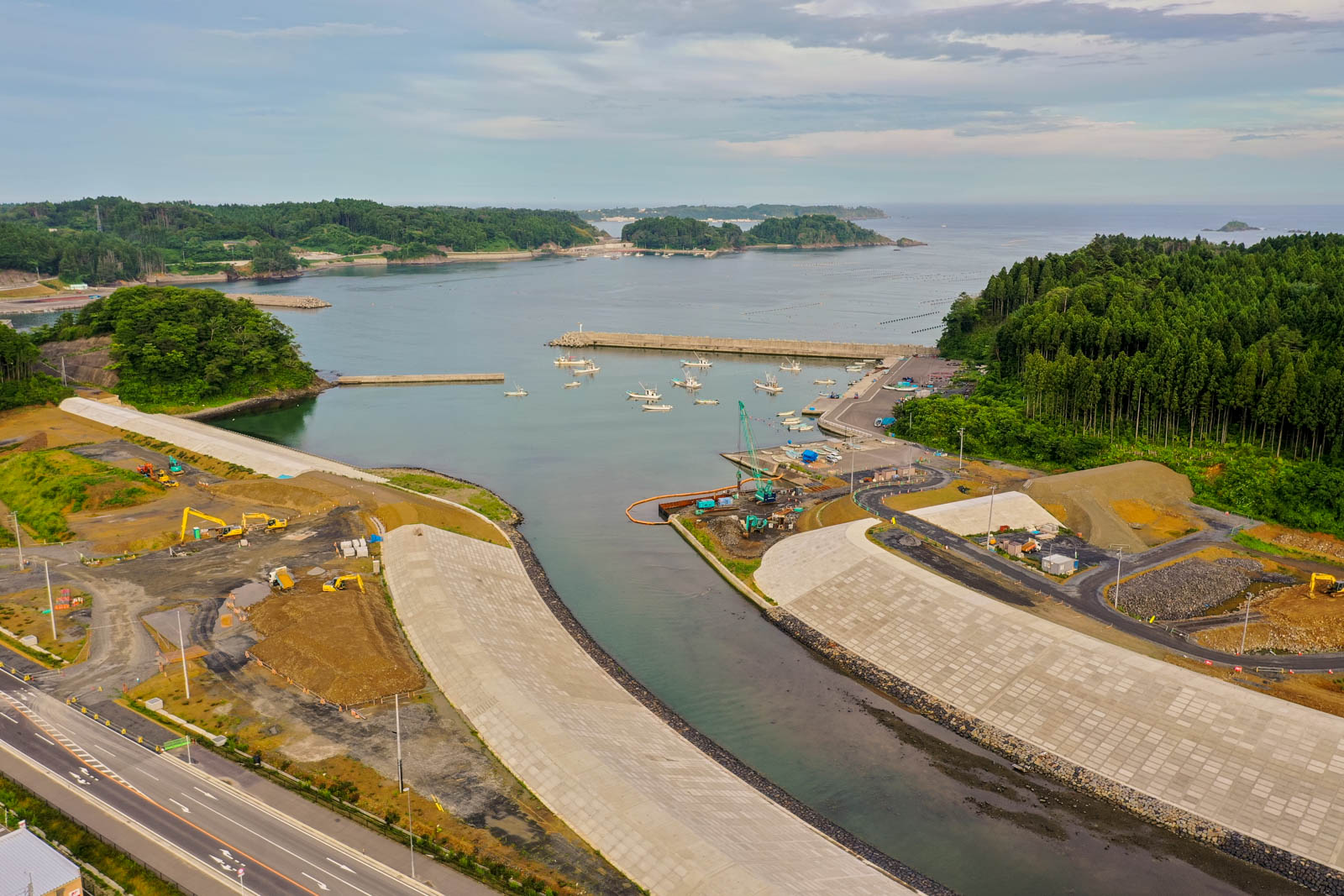 港には複数の船が停泊しており、防波堤に囲まれた海域の周囲には工事用の機材や資材が置かれ、周辺には森林や港へと続く道路が広がる、開発中の沿岸地域を空から見た写真