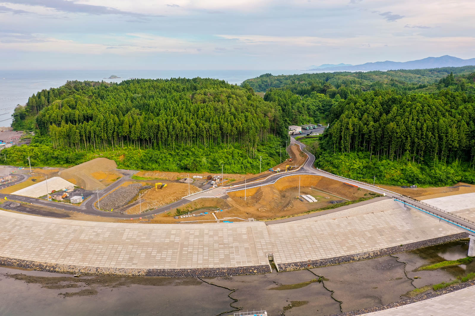 手前に大きなコンクリートの防潮堤があり、その背後には工事用の機材や資材が並ぶ建設現場が広がり、左側には海が見え、背景には緑の森林と丘が連なる沿岸地域の写真