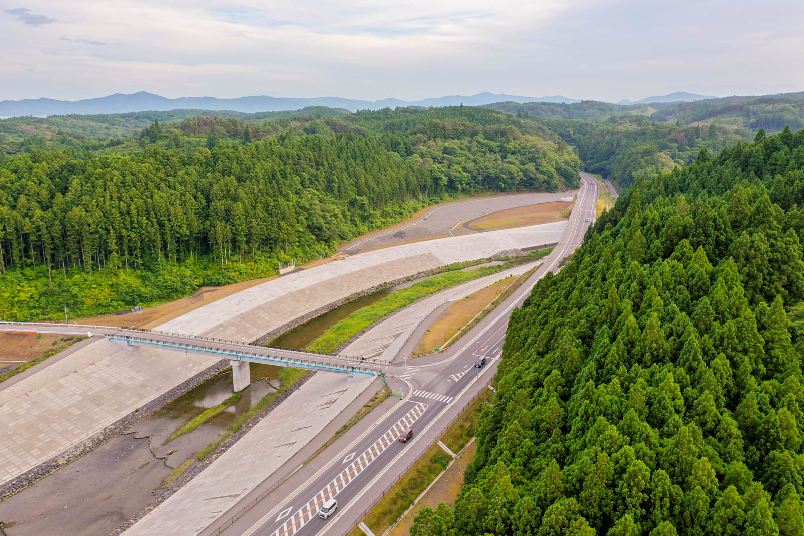 道路と橋が森林地帯を貫いており、両側には密集した緑の木々が広がり、並行して大きなコンクリート構造物が走り、背景にはなだらかな丘と遠くの山々が見える空から見た風景の写真