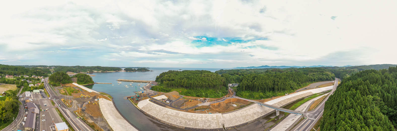 手前に大きなコンクリート構造物が並び、海や湾の周囲には緑の丘や森林が広がり、道路や歩道、工事用の車両が点在する、青空と雲が混ざる沿岸地域の写真