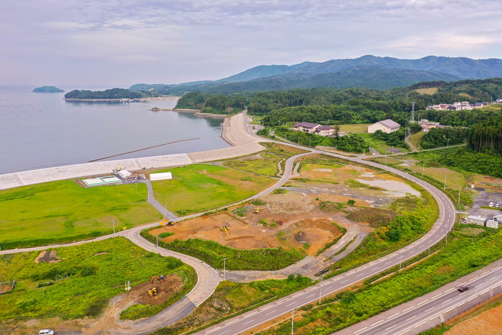 左側に広がる海と防波堤のそばに、緑の畑や工事現場、曲がりくねった道路が点在し、背景には森林に覆われた丘や山が連なる空から見た沿岸地域の写真